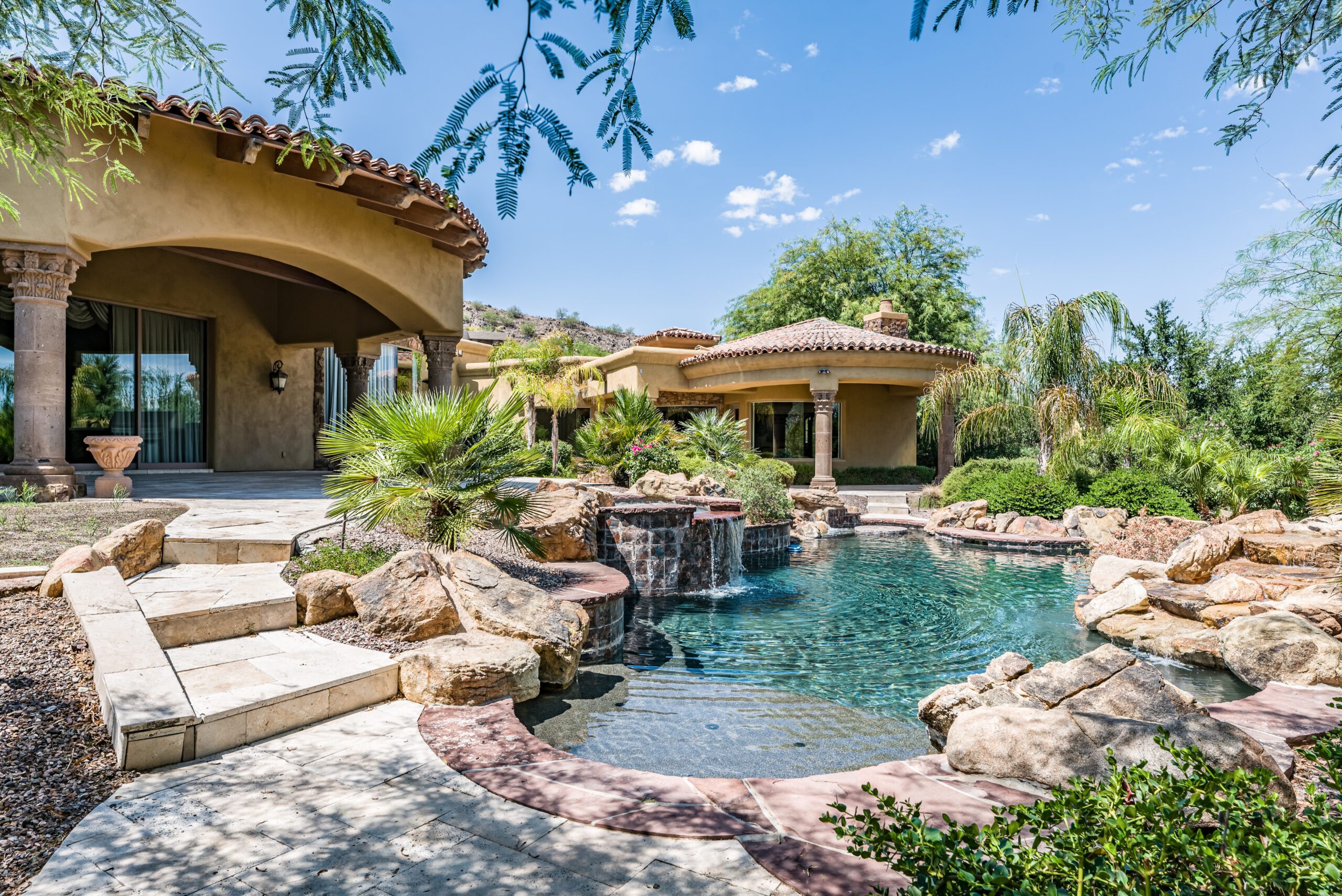 Luxury,Home,Backyard,Swimming,Pool A patio with a dining table and chairs on stone flooring, adjacent to a house with French doors. It is surrounded by lush greenery and a vibrant flower arrangement in a large pot.